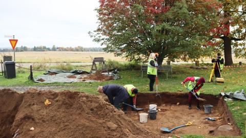 Ihmisiä arkeologisilla kaivauksia. Maahan on kaivettu kuoppa, jossa kolme henkilöä lapioi hiekkaa ämpäreihin. Maisema on syksyinen.