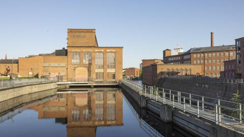 Museokeskus Vapriikki, punatiilinen kaksikerroksinen rakennus, kuvattuna ulkoa kevätiltana. Edessä patoaltaassa vettä, sivussa Tammerkoski. Taivas on pilvetön. 