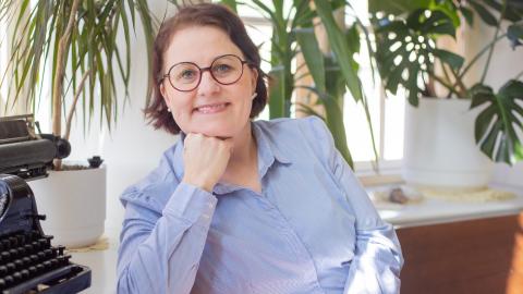Uusi nuorisopalvelujohtaja Tiina-Liisa Vehkalahti katsoo kameraan hymyillen. Ympärillä huonekasveja ja vanhanaikainen kirjoituskone.