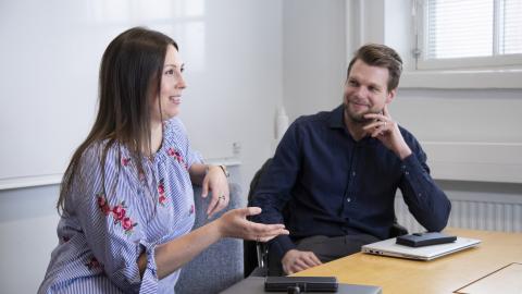 Hankintapalveluiden työntekijät Heini Kuoppamäki ja Jarkko Vesapuisto istuvat pöydän ääressä ja juttelevat.