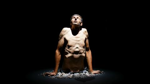 Valokuva hyperrealistisesta veistoksesta, jossa miehen ylävartalo nousee lattiasta. 