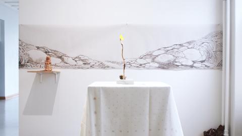 Valkoisen pöytäliinan päälle on asetetty oksannäköinen kynttilä. Seinälle on ripustettu piirustus kivikosta.