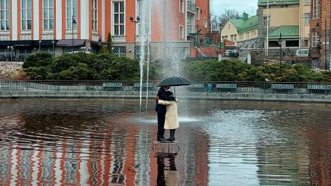 Valokuva kaupunkitilassa olevasta lammesta, jonka keskellä on vanhempaa pariskuntaa esittävä veistos. Pari seisoo sateenvarjon alla.