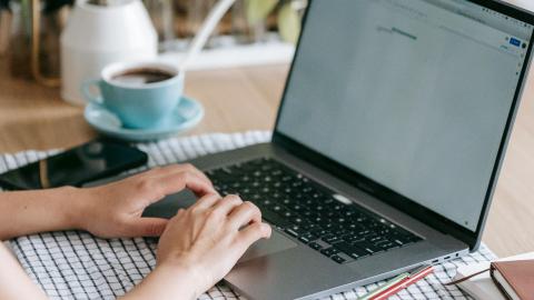 Pöydällä on kahvikuppi ja tietokone. Kädet ovat näppäimistöllä.