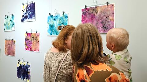 Seinällä abstrakteja vauvojen ja taaperoiden tekemiä taideteoksia. Kaksi aikuista ja toisella vauva sylissään ihastelemassa teoksia. 