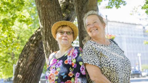 Kaksi hymyilevää naista nojaa puunrunkoon.