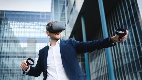 Mies seisoo modernin lasirakennuksen edessä vr-lasit päässään ja sensorit käsissään.