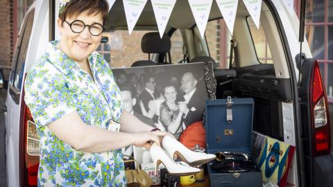 Tummahiuksinen, silmälasipäinen nainen näyttää valkoisia naisten korkokenkiä auton tavaratilan edessä.