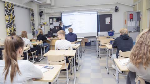 Yläasteikäisiä oppilaita koulutunnilla kuvattuna takaapäin, opettaja edessä