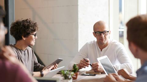 Neljä henkilöä keskustelevat kokouspöydän äärellä. Pöydällä on muistiinpanovälineitä kuten kannettavia tietokoneita, vihkoja ja kyniä. 