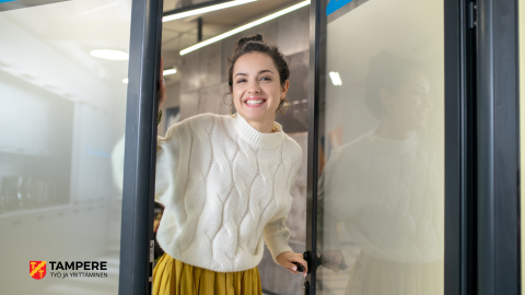 Hymyilevä nainen avaamassa ovea ja toivottamassa katsojaa tervetulleeksi.