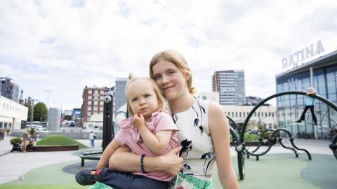 Teini-ikäinen nuori pitelee sylissään parivuotiasta sisarustaan. He istuvat Ratinan kauppakeskuksen lasten leikkipaikan luona.