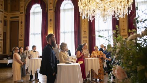 Ihmisiä suuressa salissa seisomassa pienten pyöreiden pöytien ääressä.