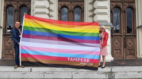 Kaksi henkilöä seisoo Tampereen Raatihuoneen rappusilla. He levittävät näkyviin suurta Tampereen omaa pride-lippua, jossa on 13 väriraitaa.