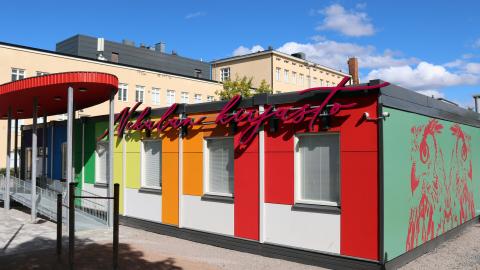 A colourful library made of Nekala containers and the oval canopy in front of it. The Nekala school building can be seen in the background. 