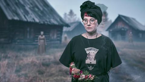 Naisella on päällään tummat vaatteeet. Kaulassa roikkuu suuri koru, jossa on mustavalkoinen kasvokuva.