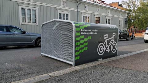 Pyörien  säilytyshangaari kadun reunassa, toisella puolella puutalo.