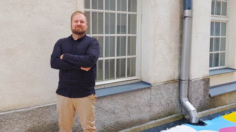 Mies seisoo kädet puuskassa vaalean rakennuksen seinämällä, taustalla ruutuikkuna.