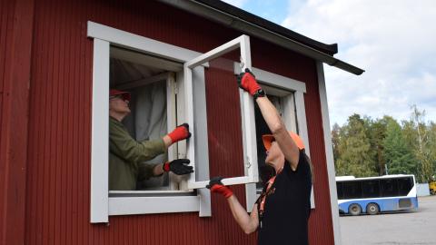 Punaisen talon ikkuna on auki ja kaksi henkilöä korjaavat sitä.