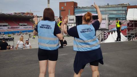 Kaksi henkilöä seisoo Ratinassa siniset liivit päällään. Liiveissä lukee lukee "miten menee".