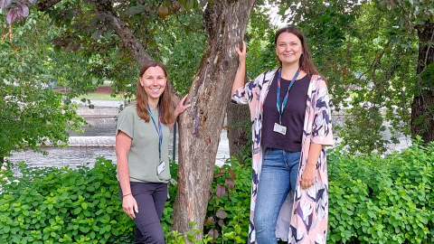 Kaksi hymyilevää naista nojaa puuhun, yksi kummallakin puolella.