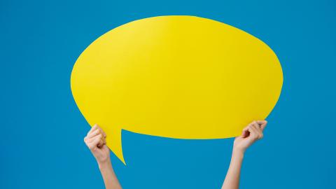 Kädet pitelevät kirkkaan keltaista puhekuplaa.