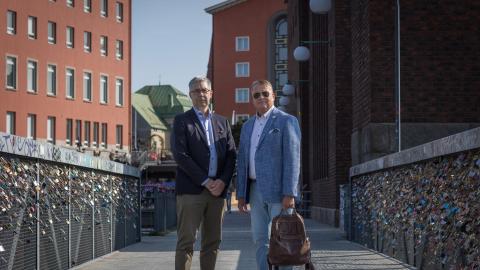 Jari Ahola ja Harri Airaksinen seisovat vierekkäin kevyenliikenteen sillalla, jonka kaiteisiin on kiinnitetty kymmeniä rakkauslukkoja, ja taustalla näkyy tiilenpunaisia korkeita rakennuksia..