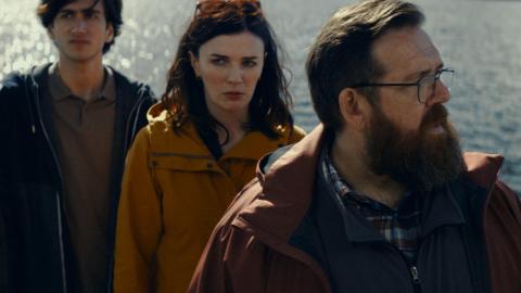 Vakavat näyttelijät seisovat laiturilla järvimaisema taustanaan.