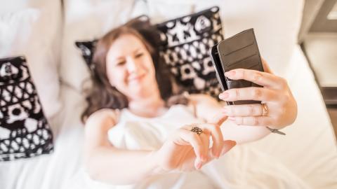 Sängyllä makoileva nainen ottaa kuvaa itsestään puhelimella.