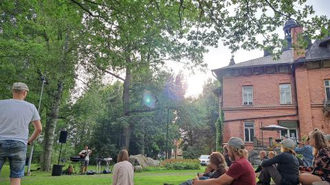 Muusikko esiintyy puistossa kesällä. Kuuntelijat istuvat nurmikolla.