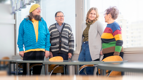 Four people are standing by the desks near the window.