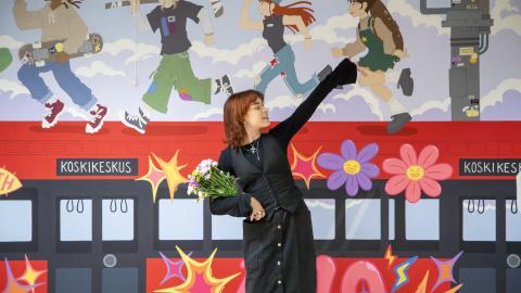 Muraalin tehnyt henkilö seisoo teoksensa edessä. Teoksessa on punainen raitiovaunu, jonka katolla juoksee nuoria. 