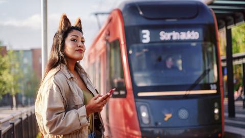 Nainen ja Ratikka ratikkapysäkillä. Naisella kännykkä kädessään ja hassut korvat päässä.