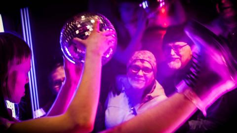 Diskopalloa käsissään pitävä hahmo esiintyy ikkunan takana kadulla seisovalle yleisölle. 