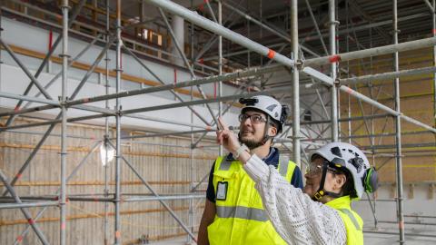 Kaksi henkilöä katselee valtuustosalin seinää. Toinen osoittaa kädellään katsesuuntaan. Kuvan tausta on täynnä rakennustelineitä.