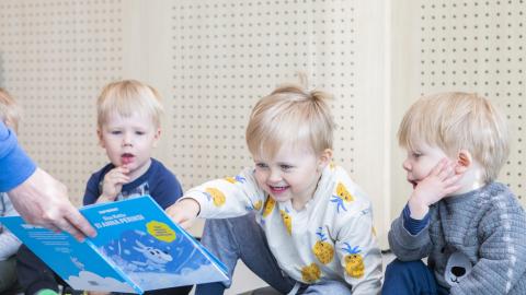 Three boys are sitting on the floor, studying a storybook that an adult is showing them.
