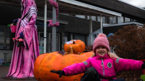 OIkealla leikkivä lapsi, vasemmalla taaempana pinkkeihin kaapuihin pukeutunut hahmo. Välissä puhallettava iso kurpitsa.