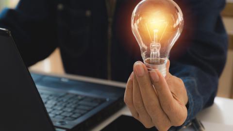 Ihminen istuu tietokoneen ääressä ja pitää kädessään hehkulamppua, jossa on valo.