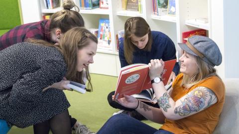 Four young people are sitting and looking at a Moomin book, with a bookshelf in the background.