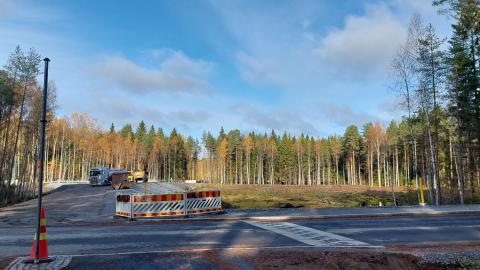 Katu, jonka takana metsän ympäröimä aukio. Keskellä katutyöpuomeja ja niiden takana kuorma-auto.