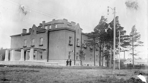 Mustavalkoisessa valokuvassa iso rakennus kuvattuna ulkoapäin. Edessä havupuita harvassa.