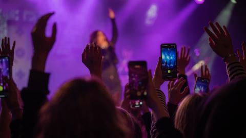 Yleisö kädet ilmassa. Etualalla puhelin, joka kuvaa lavaa, jossa näkyy esiintyjä.
