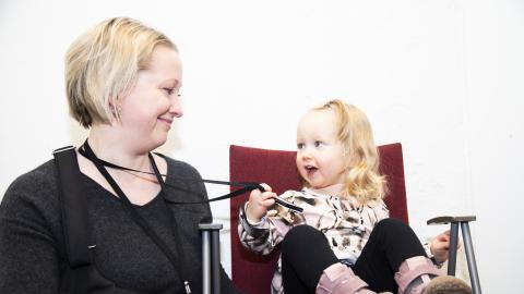 Äiti ja hänen alle kouluikäinen tyttärensä istuvat vierekkäin. He katsovat toisiaan silmiin ja hymyilevät. Tyttö pitää kädessään äidin kaulassa olevaa kulkulupakorttia.