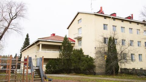 Rakennus on vaalean keltainen kolmikerroksinen ja siinä on matala siipirakennus, pihassa lasten lasten leikkipaikka ja ympärillä puita.