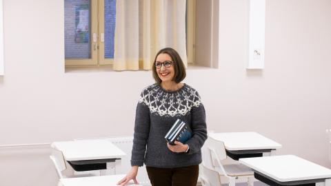 The teacher stands in the classroom in the middle of the desks, books in hand.