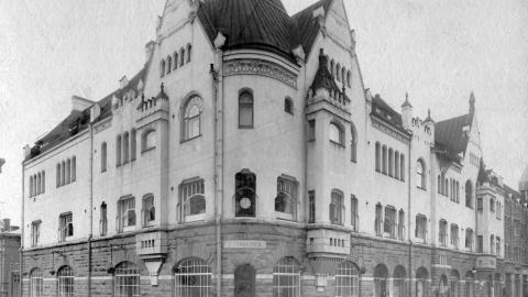 Mustavalkoisessa valokuvassa iso nelikerroksinen rakennus, jonka alatasossa on liiketilaa.