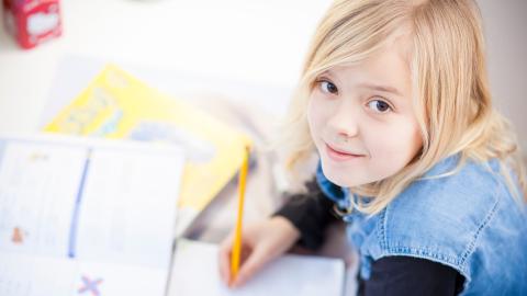 Vaaleahiuksinen lapsi on tekemässä läksyjä koulupöydän ääressä. Hän on nostanut katseensa kohti katsojaa.