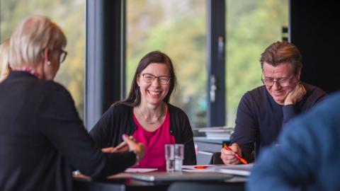 Hymyileviä ihmisiä istuu pöydän ääressä. Pöydällä tietokone ja papereita.