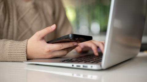 A person's hands, one on a laptop keyboard and the other holding a smartphone.