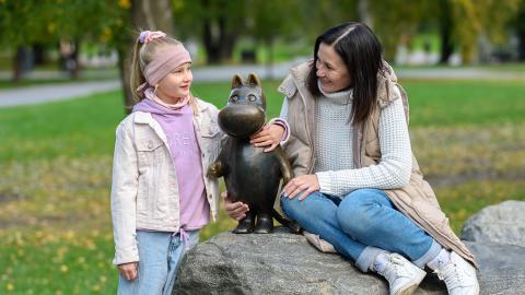 A blonde girl and a dark-haired woman gathered around a statue of the Moomintroll.